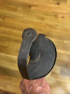 Close-up of a dark, worn metal tool with a circular disk and curved lever being held by a hand, wooden floor in the background.