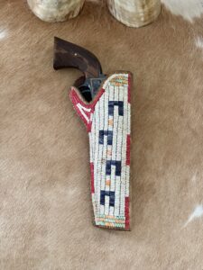 A revolver with a wooden grip rests in a decorative beaded leather holster on a brown hide surface.